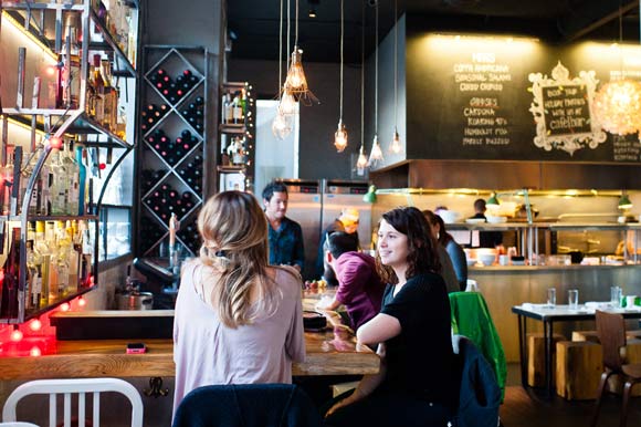 People enjoy drinks at the bar, while an open kitchen is featured in the background at Cafébar in West Washington Park. 